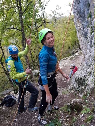 Trois personnes font de l'escalade en plein air. Une femme souriante en tenue d'escalade se tient au premier plan, tandis que deux autres personnes l'assistent près d'une falaise rocheuse et d'arbres.