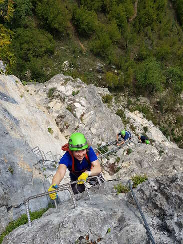 Trois grimpeurs escaladent une falaise rocheuse abrupte à l'aide d'échelons métalliques et d'équipements de sécurité. Ils portent des casques, des gants et des harnais. Une forêt verdoyante s'étend en contrebas.