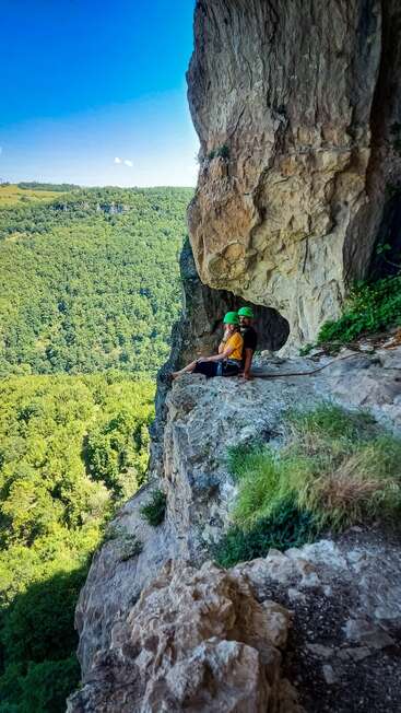 Deux alpinistes, portant des casques verts, sont assis sur le rebord d'une falaise rocheuse. Derrière eux se trouve une vaste forêt verte et, au-dessus, un ciel bleu vif sans nuages.