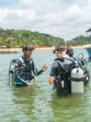 Three people wearing scuba diving gear are standing in shallow water. One appears to be instructing the others, with a lush, green coastline visible behind them.