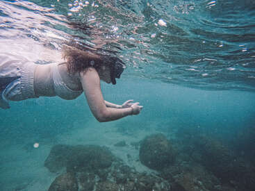 A person in a white swimsuit snorkels underwater, surrounded by clear blue water and rocks below, exploring the tranquil marine environment with curiosity and focus.