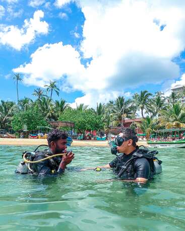 Two men in scuba gear stand in clear water near a tropical beach. Palm trees and beach structures are visible under a bright blue, partly cloudy sky.