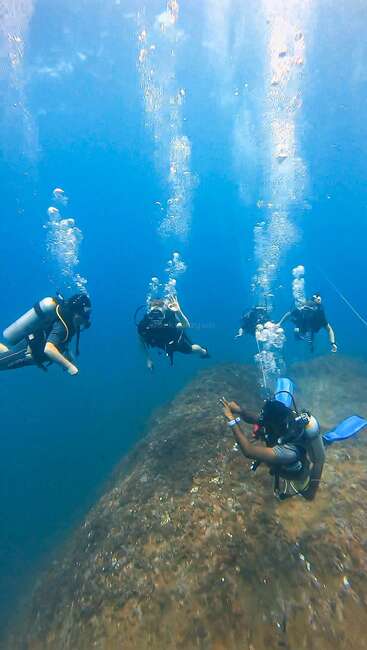 Five scuba divers explore underwater, surrounded by bubbles rising to the surface. One diver gestures, while others float above sandy terrain, enjoying the aquatic adventure together.