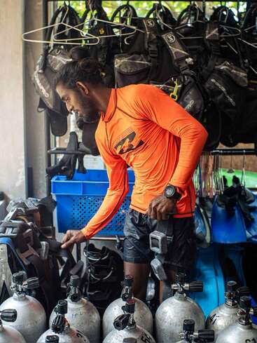 A man in an orange rash guard prepares scuba diving gear, surrounded by air tanks, weight belts, fins, and wetsuits in a well-organized dive shop.