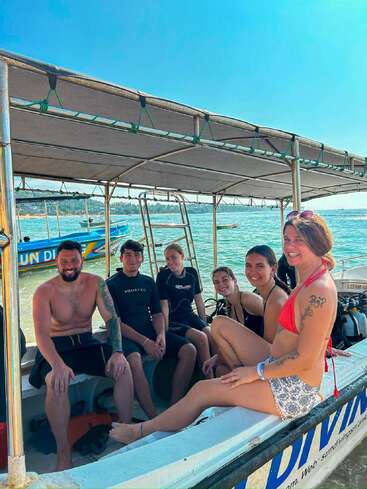 Six people sit smiling on a boat, dressed in swimwear and wetsuits, enjoying sunny weather by the water, ready for a fun diving adventure together.