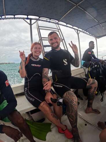 Two people in wetsuits are sitting on a boat, smiling and making peace signs. Other divers are nearby. The atmosphere is cheerful, adventurous, and relaxed.