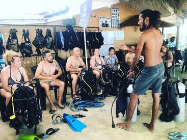 The image depicts a group of people gathered in a room, likely a scuba diving shop, with a man standing and addressing the group.