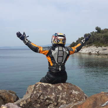 A person wearing a helmet and jacket sits on a rock by the water with their arms outstretched, facing away from the camera.