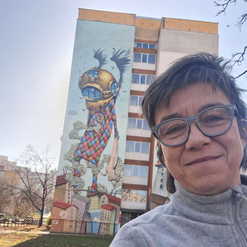 The image depicts a woman standing in front of a tall building featuring a vibrant mural of a person wearing a helmet and colorful clothing, set against a serene outdoor backdrop.