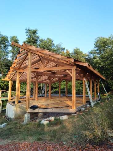 A timber frame structure under construction stands on a grassy plot. Wooden beams form the frame and roof, surrounded by trees, ladders, and construction materials. Clear sky above.