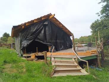 This image shows a rustic wooden structure with a sloped roof, partially covered by black tarp. Wooden stairs lead up to a deck surrounded by greenery.
