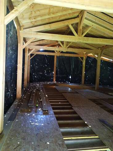 This image shows the wooden frame of a building under construction, with beams and unfinished flooring. Sunlight shines through small holes in the dark plastic sheeting.