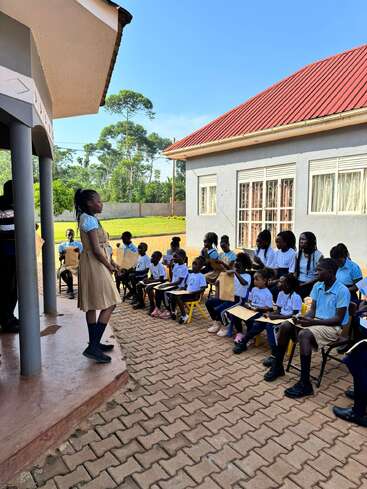 L'image représente une jeune fille debout sous un porche, s'adressant à un groupe d'enfants assis à l'extérieur d'un bâtiment au toit rouge, probablement engagés dans une activité éducative en plein air.