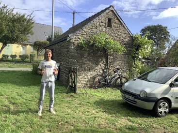 A young man in a white Puma shirt stands on green grass, near a stone shed, with two bicycles and a silver car beside him, on a sunny day.