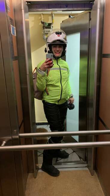 Une personne portant un casque, une veste vert fluo et un pantalon noir prend un selfie dans un ascenseur, tenant un téléphone et un petit sac noir.