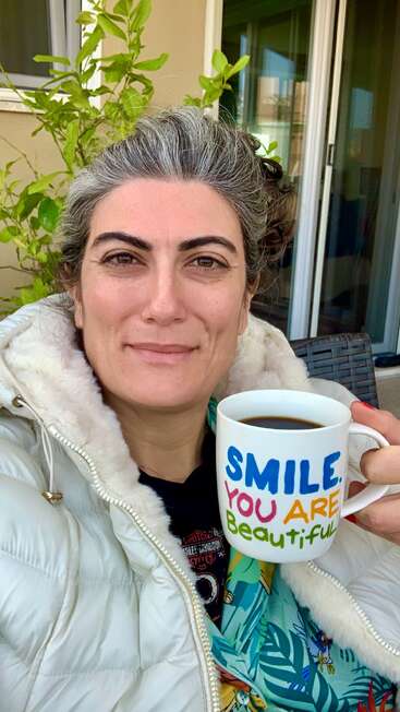Une femme aux cheveux poivre et sel sourit, tenant une tasse à café avec un texte coloré : "SMILE. YOU ARE Beautiful" (Souriez. Vous êtes belle). Elle porte une veste blanche confortable à l'extérieur.