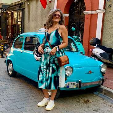 Une femme souriante, vêtue d'une élégante robe bleue, se tient devant une voiture turquoise d'époque, un casque à la main, dans une rue animée de la ville, avec des scooters à proximité.