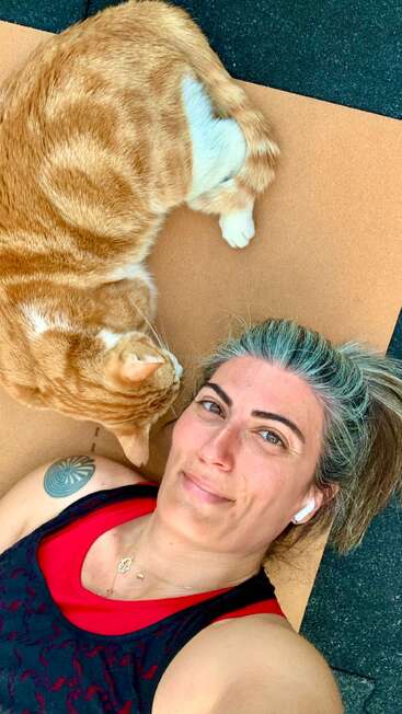 Une femme aux cheveux gris striés est allongée sur un tapis à côté d'un chat orange recroquevillé. Elle sourit, portant des écouteurs et un haut noir avec un tatouage visible.