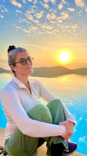 Une femme assise paisiblement au bord d'une piscine au lever ou au coucher du soleil, portant des lunettes, une veste blanche zippée et un pantalon vert. Atmosphère calme et réfléchie avec un ciel magnifique.