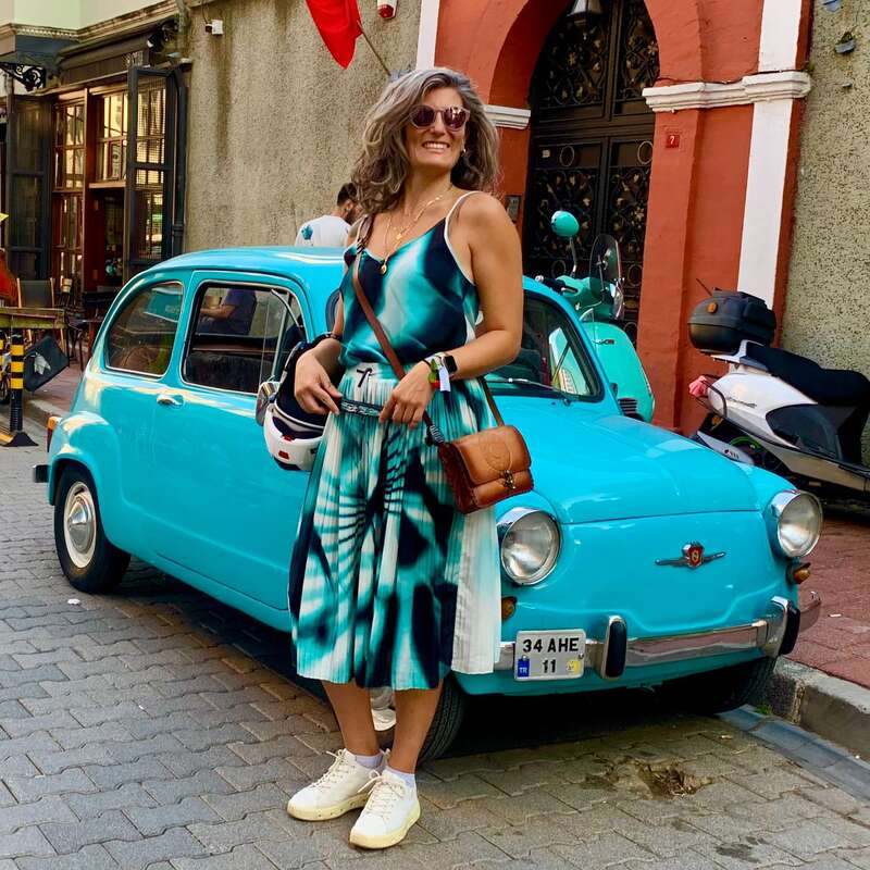 Une femme souriante, vêtue d'une élégante robe bleue, se tient devant une voiture turquoise d'époque, un casque à la main, dans une rue animée de la ville, avec des scooters à proximité.
