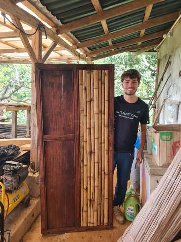 Un homme souriant se tient à côté d'une armoire en bois faite main avec des accents de bambou, sous un toit rustique. L'atelier est entouré de matériaux naturels, d'outils et de verdure.