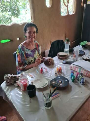 Une femme souriante est assise à une table, en train de peindre un objet rond. Des fournitures artistiques, des pinceaux, des peintures et des pierres décorées l'entourent dans un cadre artistique et chaleureux.