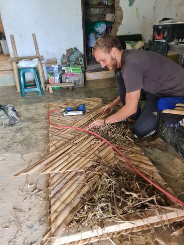 Un homme fabrique des objets en bambou et en paille dans une pièce rustique. Il semble concentré et heureux, entouré de divers outils, fournitures et matériaux de construction.