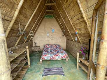 Cette image montre une cabane confortable en bambou avec un lit coloré, des capteurs de rêves accrochés au mur, des étagères, une petite table, des plantes en pot et une décoration naturelle.