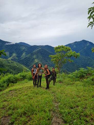 Quatre personnes souriantes se tiennent debout au sommet d'une colline verdoyante, entourées de montagnes luxuriantes et d'un ciel nuageux, profitant de la nature et d'une randonnée aventureuse en pleine nature.