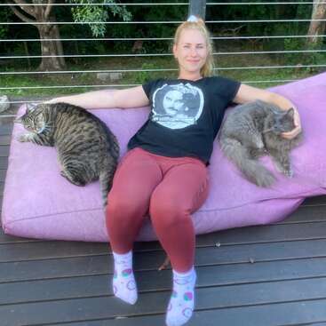 A woman sits comfortably on a pink outdoor cushion, smiling, with a tabby cat on her left and a fluffy gray cat on her right.
