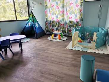 Das Bild zeigt ein Spielzimmer mit einem Tipi, einem Laufstall und einem Tisch. Ein tropischer Vorhang, ein Holzboden und hellblaue Wände schaffen eine gemütliche Atmosphäre für Kinder zum Spielen.