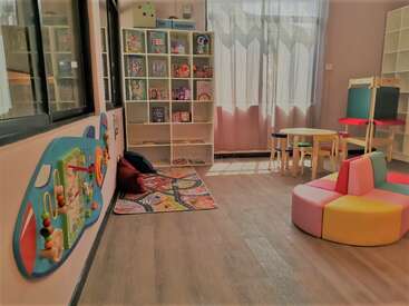 Das Bild zeigt ein gut beleuchtetes, organisiertes Spielzimmer mit einer Vielzahl von Spielsachen und Möbeln, darunter ein Bücherregal, ein Tisch, Stühle und bunte Sitzgelegenheiten, die eine einladende Umgebung schaffen.
