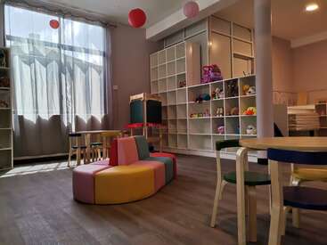 Das Bild zeigt ein gut beleuchtetes Spielzimmer mit einem Fenster, Regalen und farbenfrohen Möbeln mit einem Tisch und Stühlen sowie einer Vielzahl von Spielzeugen und Stofftieren.