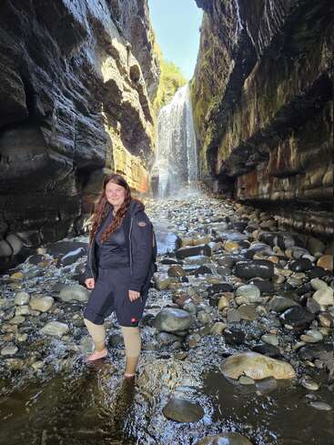 Uma mulher está descalça sobre as rochas em um desfiladeiro estreito com um riacho. A luz do sol ilumina uma linda cachoeira ao fundo, cercada por penhascos cobertos de musgo.