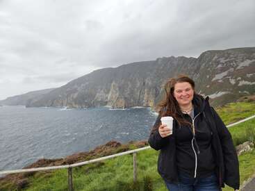 Uma mulher está de pé, sorrindo, segurando uma xícara de café, com penhascos escarpados e o oceano ao fundo. Ela está vestindo uma jaqueta escura, e o céu está nublado.