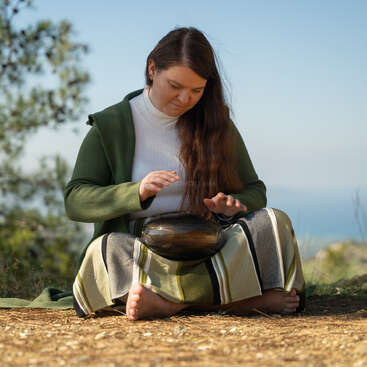 Une femme est assise en tailleur à l'extérieur et joue du handpan. Elle porte une veste verte et un pantalon rayé. Des arbres et un ciel magnifique composent le décor naturel.