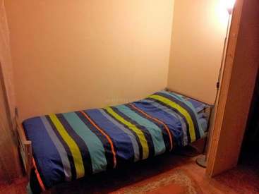 A imagem mostra um quarto pequeno e mal iluminado com uma cama de solteiro com um edredom listrado, uma luminária de chão e uma porta que leva a outro quarto.