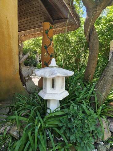 A white stone lantern stands amidst lush green foliage near a wooden pillar with floral designs, under a rustic wooden roof in a serene garden setting.