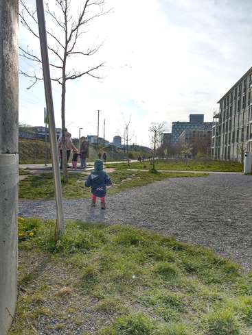 La imagen muestra a un niño caminando por un sendero de grava, rodeado de hierba y árboles, con un paisaje urbano al fondo y gente de pie cerca.