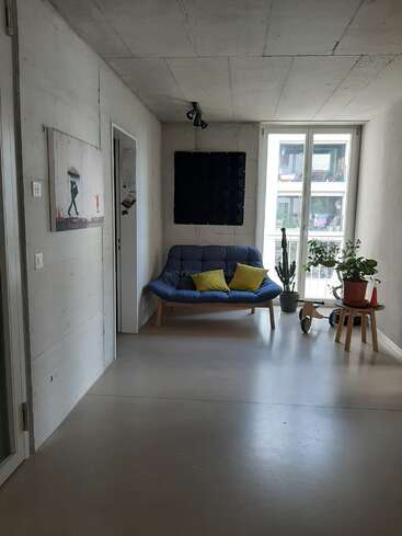 La imagen muestra una habitación moderna de paredes y techo blancos, con un sofá futón azul, un cuadro y una ventana con vistas al exterior.