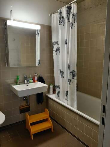 La imagen muestra un cuarto de baño con lavabo, espejo y bañera blancos, con una cortina de ducha con estampado de cebra y un taburete amarillo en la esquina.