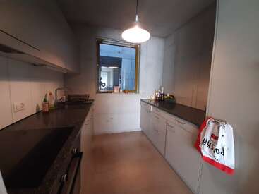 La imagen muestra una cocina compacta con armarios blancos, encimeras negras y una ventana, en la que aparece una toalla colgante con la palabra "POSKA" y un borde rojo.