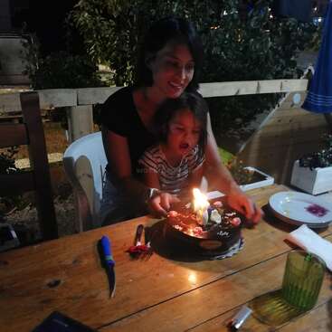 Das Bild zeigt eine Frau und ein Kind, die an einem Holztisch sitzen und mit einem mit Kerzen verzierten Kuchen vor einem ruhigen Hintergrund im Freien feiern.