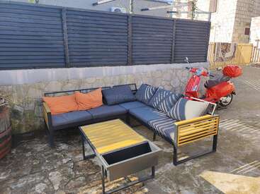Sitzbereich im Freien mit einem Ecksofa, gestreiften und orangefarbenen Kissen, einem Couchtisch aus Holz und einem roten Roller, der daneben geparkt ist, vor einer Steinmauer und einem Zaun.