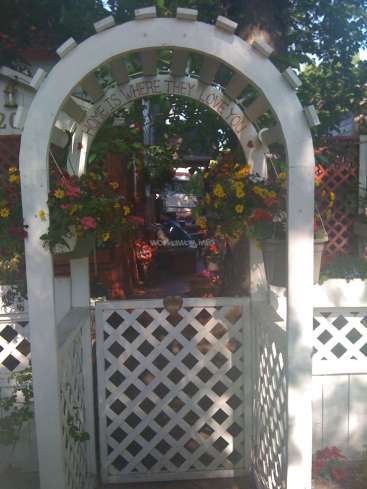 The image depicts a white arched gate with a lattice door, adorned with flowers and a sign that reads "HOME IS WHERE THEY LOVE YOU".