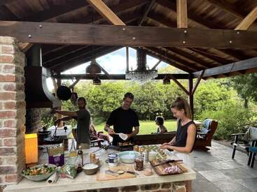 Three people cook together in an outdoor kitchen under a wooden pavilion. Fresh ingredients and prepared dishes cover the counter, while others relax outside enjoying nature.