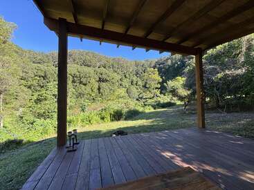 A wooden porch overlooks a lush, green forested hillside under a clear blue sky, creating a peaceful, inviting outdoor setting with nature all around.
