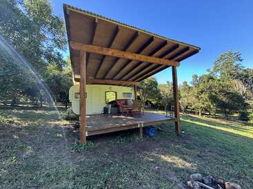 A cozy caravan sits on a wooden deck with a large slanted roof, surrounded by green grass and trees, under a clear, bright blue sky.