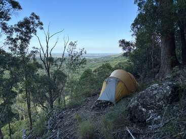 A yellow tent is pitched on a rocky, forested hillside overlooking a lush green valley with trees, under a clear blue sky, peaceful and remote.