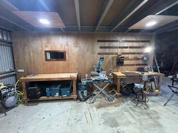 This image shows a well-organized woodworking workshop with wooden benches, various power tools, equipment, a wooden wall, pegboard system, and overhead lighting in a spacious area.